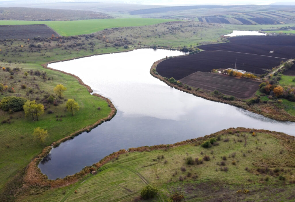 Aerial drone view of nature in Moldova, lake with cloudy sky reflecting, sown fileds, trees, hills on the background