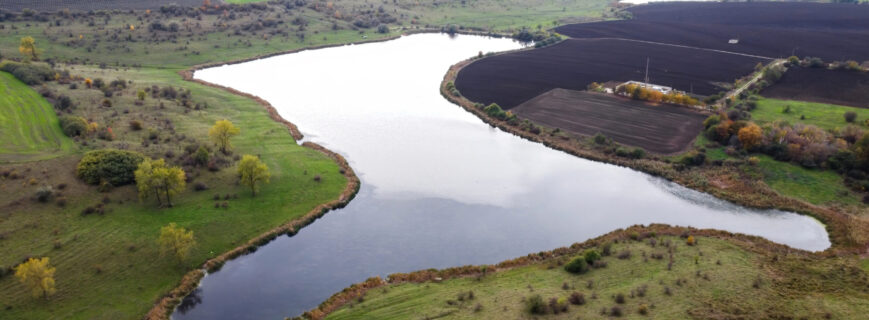 Aerial drone view of nature in Moldova, lake with cloudy sky reflecting, sown fileds, trees, hills on the background